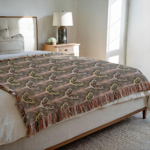 A woven cotton throw featuring an intricate luna moth pattern in bronze and muted earth tones, framed by soft fringe.